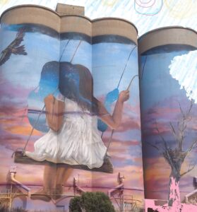 On tall cement wheat silos there is a painted mural of a girl on a swing facing away from the viewer, showing a shadow image behind the girl to suggest the swing is in motion. In the background of blue sky and ochre landscape there is a wedge tailed eagle in flight.